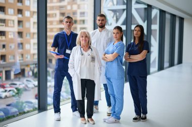 Sağlık uzmanları gösterişli bir hastane koridorunda güvenle duruyorlar. Önlük ve laboratuvar önlüğü giyiyorlar. Takım çalışması ve hasta bakımına bağlılıklarını sergiliyorlar..