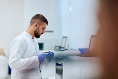 Beyaz laboratuvar önlüklü bir bilim adamı, gündüz saatlerinde gelişmiş ekipman ve aydınlatmalarla dolu modern bir laboratuvardaki bir aleti kullanarak işine odaklanmış durumda..