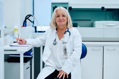 Bir tıp uzmanı parlak bir ofiste rahat bir şekilde oturur ve bir masada notlar alır. Odada, hastalar için hoş geldin ortamı yansıtan tıbbi araç ve gereçler bulunuyor..