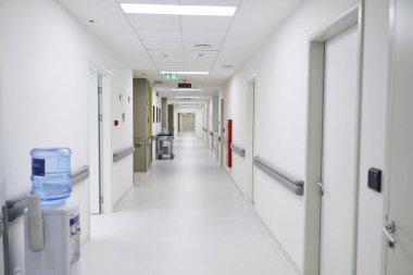 Parlak bir hastane koridorunda temiz, beyaz duvarlar ve yerler var. Sağlık hizmetlerinin iyi korunduğunu gösteren tıbbi ekipmanlar su dağıtıcısının yanında görülebiliyor..