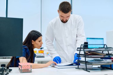 İki bilimadamı, parlak ve modern bir laboratuvarda çalışma masasında araştırma belgelerini gözden geçiriyorlar. Kadın odaklanırken, erkek sayfalara bakar..