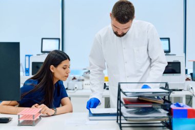 İki sağlık uzmanı laboratuvar masasında sonuçları tartışıp analiz ediyorlar. Çevrede gelişmiş tıbbi ekipmanlar ve temiz bir çalışma alanı bulunmaktadır..