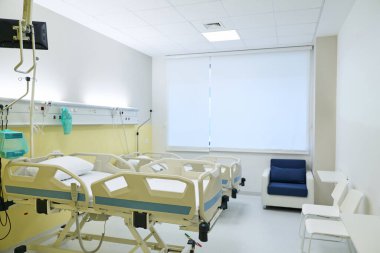 Geniş hastane odasında birkaç hasta yatağı ve tıbbi gereçler var. Ziyaretçiler için oturma alanı ve doğal ışığa izin veren bir pencere mevcuttur..