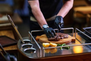 Siyah eldiven giyen bir şef zarif, profesyonel bir mutfakta ahşap bir kesme tahtasının üzerinde biftek dilimliyor. Çevredeki limonlar ve yeşiller lezzetli, ev yapımı bir akşam yemeği hazırlıyor..