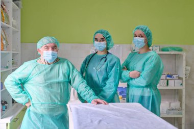 Koruyucu elbiseler, maskeler ve kepler giyen profesyonel bir tıbbi ekip, temiz bir ameliyathanede bir hastanın yanında duruyor. Güvenli hasta bakımı için gerekli güven, odaklanma ve takım çalışmasını yansıtıyorlar..
