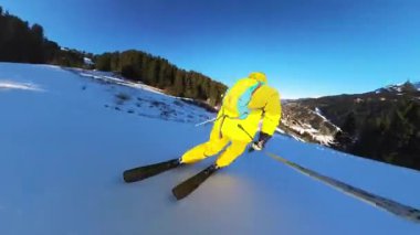Bir kayakçının el değmemiş, güneşten sırılsıklam olmuş bir dağ yamacını oymasıyla birinci şahıs bakış açısının heyecanını yaşayın. Parlak sarı kayak takımı beyaz kara ve açık mavi gökyüzüne karşı duruyor.