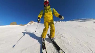 POV görüntülerinde parlak sarı bir kayakçının el değmemiş karla kaplı bir dağda ustalıkla gezinerek parlak mavi gökyüzü ve sıcak güneş altında toz izleri bırakması görülüyor..