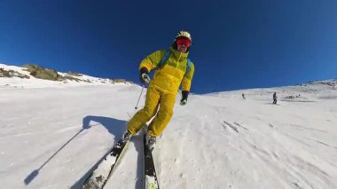 Sarı kayak kıyafeti giymiş bir adam temiz mavi bir gökyüzünün altında el değmemiş bir dağ yamacında kayak yapmaktan hoşlanır. Kar tazedir ve güneş ışıl ışıl parlar. İdeal bir kış sporu deneyimi yaratır..