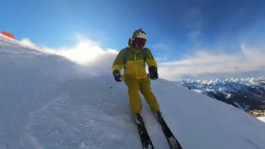 Parlak, güneşli bir kış gününde yokuş aşağı kaymanın heyecanını yaşayın. Sarı kayak kıyafetli bir insan güzel bir dağ yamacındaki taze karların arasından zarif bir şekilde geçerek özgürlüğün tadını çıkarır.