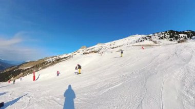 İnsanlar açık mavi bir gökyüzünün altındaki geniş, karlı bir dağ yamacında kayak ve snowboard yapmayı severler. Uçsuz bucaksız vadi ve uzak tepelerin panoramik manzarası sakin bir kış sporu sahnesi yaratır..