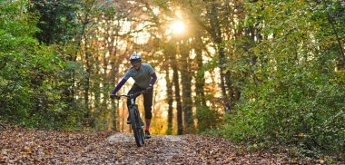 Odaklanmış bir bisikletçi, sonbahar boyunca yapraklarla kaplı bir orman patikası boyunca dağ bisikleti sürüyor ve güneş ağaçların arasından gözetliyor. Sahne, açık hava fitness, doğa ve maceranın heyecanını yakalar..