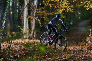 Kask takan bir bisikletçi, dağ bisikleti üzerinde yapraklarla kaplı bir orman yolunda sonbahar rengini, odak noktasını ve açık hava macerasını dinamik ve manzaralı bir yolculukta yakalar..