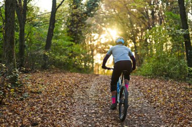 Bir bisikletçi günbatımında yapraklarla kaplı orman patikası boyunca gezer, açık hava fitnesinin ve sonbahar doğasının dinginliğinin tadını çıkarır..