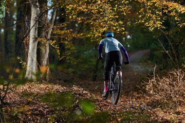 Kasklı ve koruyucu teçhizatlı bir bisikletçi düşerken yapraklarla kaplı orman yolunda dağ bisikleti sürüyor. Sahne, açık hava sporunu, odaklanmayı ve sonbahar renklerinin güzelliğini yakalıyor..