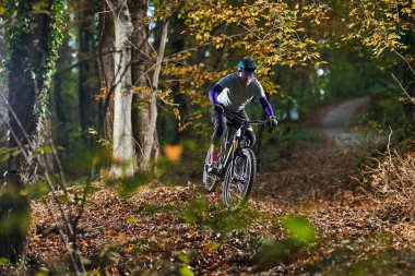 Kask takan bir dağ bisikletçisi sonbaharda yapraklarla kaplı bir orman patikasında gezer. Dinamik hareket, canlı yeşillik, ve engebeli arazi açık hava macerası ve zindelik iletin.
