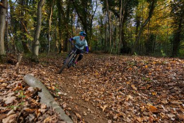 Bir bisikletçi sonbaharda yapraklarla kaplı bir orman patikasında dağ bisikleti sürüyor. Sürücü, uzun ağaçlar ve yumuşak ışıklar arasında keskin bir dönüş yapmaya odaklanmış bir kask ve koruyucu kıyafet giyer..