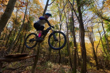 Sonbahar ormanı sahnesinde dağ bisikleti süren bir binici kütük engelinin üzerinden atlıyor. Zengin sonbahar renkleri, uzun ağaçlar ve yüksek enerjili açık hava macerası hissi..
