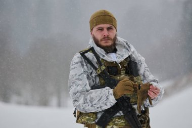 Beyaz kış kamuflajlı ve taktik teçhizatlı sakallı bir asker soğuk bir orman devriyesi sırasında kar altında dikilirken eldiven ve tüfek tutuyor ihtiyatı, dayanıklılığı ve hazırlığı iletiyor..