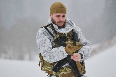 Beyaz kış kamuflajı giyen sakallı bir asker soğuk hava eğitimi sırasında yağan karda dururken taktik eldivenlerini ayarlar. Görünür savaş yeleği, tüfek ve odaklanmış ifade aktarımı