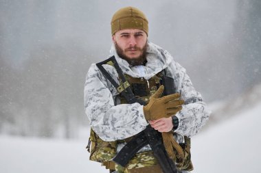 Beyaz kışlık kamuflaj ve taktik teçhizat takan uyanık bir asker, ağır kar yağışı altında durup, tüfek taşırken eldivenlerini ayarlıyor. Soğuk, odaklanmış ve uzak bir yerdeki kış operasyonlarına hazır.