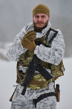 Beyaz kış kamuflajı ve taktik yelek giymiş bir asker kar yağarken elinde tüfekle eldivenli elleri ısıtıyor. Dayanıklı, odaklanmış ifade hazır olmayı, hayatta kalmayı ve soğuk havayı iletir.