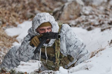 Beyaz kış kamuflajı giyen bir asker, engebeli bir arazide soğuk hava devriyesi sırasında odaklanmış ve tetikte olan karlı bir siperden hafif kar yağışı ve kışın seyrek bitki örtüsü ile bir tüfeği hedefler..