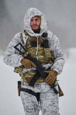 Beyaz kışlık kamuflaj ve taktik yelek giyen silahlı bir asker, soğuk hava devriyesi veya eğitim tatbikatında kar yağarken elinde bir tüfek tutuyor.