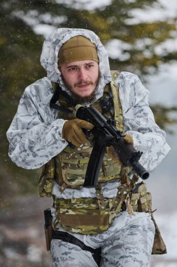 Beyaz kar kamuflajı ve taktik yelek giyen silahlı asker, soğuk orman koşullarında bir saldırı tüfeği taşırken tetikte ve odaklanmış kış devriyesinde kar altında hareket ediyor..