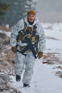Beyaz kış kamuflajı ve taktiksel yelek giyen bir asker, soğuk, sert ve karlı koşullarda ilerlerken odak ve dayanıklılık gösteren bir tüfekle karla kaplı bir orman yolunda devriye geziyor..
