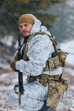 Beyaz kışlık kamuflaj ve taktik teçhizat giyen silahlı asker, kar altında diz çöküp, elinde tüfekle ormanı tarıyor. Huysuz, odaklanmış bir sahne. Hazırlık, hayatta kalma ve askeriyeyi gösteriyor.