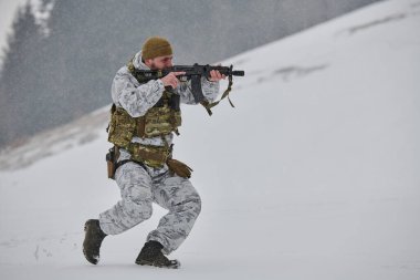 Kış kamuflajı ve taktik yelek giyen asker, karlı bir ormanda soğuk hava tatbikatı sırasında bir tüfeği nişan alırken kar yağışı içinde koşuyor..