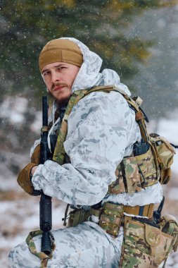Beyaz kış kamuflajı ve taktik yelek giymiş bir asker, karlı bir ormanda tetikte beklerken bir tüfek tutuyor ve soğuk hava operasyonları sırasında hazırlık, hayatta kalma ve dayanıklılık taşıyor..