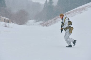 Beyaz kış kamuflajlı bir asker hafif kar yağışı sırasında karlı bir vadide taktik teçhizat ve bir tüfek taşıyarak ilerler. Sahne soğuk, hazır, eğitimli ve yalnız hareket eder