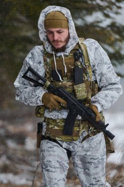 Beyaz kışlık kamuflaj ve taktik teçhizat kuşanmış silahlı askerler karlı bir ormanda devriye geziyor ve elinde bir saldırı tüfeği var. Odaklanmış ifade, soğuk hava ve göreve hazır duruş ihtiyatı iletir.
