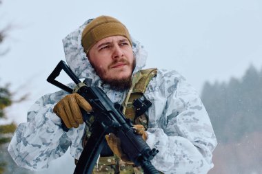 Beyaz kışlık kamuflaj ve taktik teçhizat kuşanmış silahlı asker, karlı bir orman manzarasını tararken bir tüfek tutuyor, ihtiyatı, hazırlığı ve soğuk hava askeri operasyonlarını iletiyor..