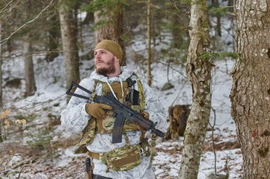 Kışın kamuflaj yapan bir asker, karlı bir ormanda, bir saldırı tüfeği ile devriye geziyor ve bir soğuk hava tatbikatı sırasında çevreyi tararken taktik ekipmanlar takıyor..