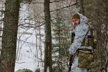 Beyaz kış kamuflajı giyen ve taktik teçhizatı olan bir asker karlı bir ormanda devriye geziyor, tüfeği kavrıyor ve omzunun üzerinden bakıyor. Soğuk atmosfer gerginlik, hazırlık, hayatta kalma ve