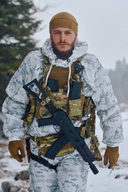 Beyaz kış kamuflajı ve taktiksel yelek giyen silahlı bir asker karlı bir ormanda elinde bir tüfek ve teçhizatla yürüyor. Görüntü hazırlığı, dayanıklılığı ve soğuk hava askeriyesini gösteriyor.