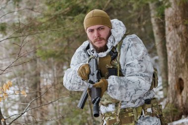 Beyaz kışlık kamuflaj ve taktik teçhizat kuşanmış silahlı bir asker karlı bir ormanda yürürken bir tüfek tutuyor. Soğuk hava devriyesi sırasında tetikte, hayatta kalma ve askeri hazırlık durumu bildiriyor..