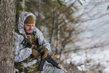 Beyaz kış kamuflajlı bir asker, soğuk hava eğitim devriyesi sırasında elinde tüfekle bir ağaca yaslanmış, kar ve orman örtüsü altında ihtiyatı, dayanıklılığı ve dikkati elden bırakmıyor..