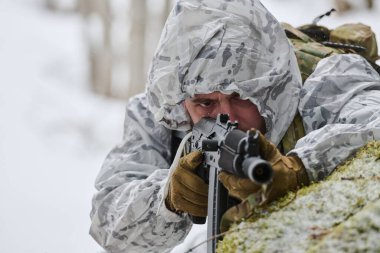 Beyaz kış kamuflajı ve eldivenleri giyen silahlı asker, soğuk hava askeri eğitimi sırasında veya karlı bir havada taktik devriye sırasında odaklanmış bir tüfeği hedefleyen yosunlu kar örtüsüne yatar.