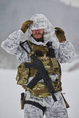Beyaz kış kamuflajı giyen bir asker, karlı, dağlık bir eğitimde veya devriyede soğuk hava operasyonları sırasında taktik yelek, tüfek ve eldiven ile donatılmış kar yağışı altında başlığını düzeltir.