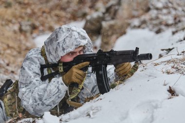 Beyaz kış kamuflajlı bir asker karlı bir siperde yatıyor ve soğuk hava eğitimi sırasında bir saldırı tüfeğini hedef alıyor. Odaklanmış ve gizlenmiş bir şekilde, engebeli araziye karışıyor. Etrafında kar yağıyor..