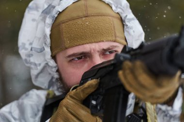Beyaz kış kamuflajlı bir askerin karlı bir tatbikat sırasında tüfeği hedeflediği yakın plan. Taktik eldiven ve teçhizatla odaklanmış, kararlı ifade, konsantrasyon ve soğuk hava iletimi