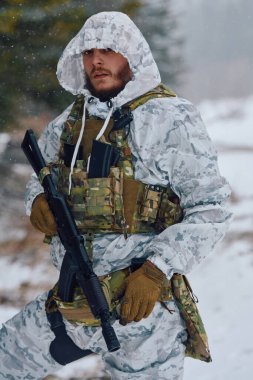 Beyaz kış kamuflajı ve taktik yelek giyen bir erkek asker, karlı bir ormanda durup hazırlık, hayatta kalma, soğuk hava operasyonları ve kışın askeri devriye görevleri yaparken tüfeğini tutuyor.