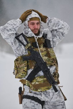 Beyaz kar kamuflajı ve taktik yelek giyen kış askeri, soğuk hava eğitimi sırasında yağan karda dikilirken başlığı düzeltiyor. Odaklanma, yorgunluk ve askeri alanın yoğunluğunu aktarıyor.