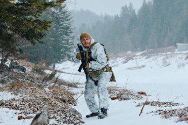 Silahlı asker kış devriyesinde karlı bir orman boyunca beyaz kamuflaj ve taktik teçhizat giyerek ihtiyatlı bir şekilde ilerliyor. Atmosferik kar yağışı ve yoğun çam ağaçları gergin, hayatta kalmaya odaklı bir sahne yaratır..