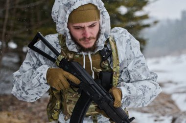Beyaz kış kamuflajı ve taktik teçhizatı olan bir asker soğuk hava askeri eğitimi sırasında karlı bir ormanda dikilirken tüfeğini ayarlıyor.