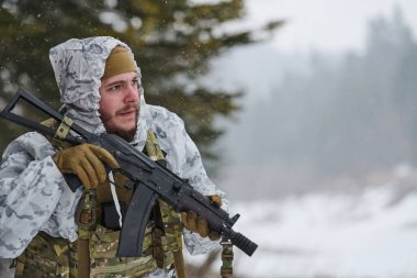 Beyaz kış kamuflajı ve taktik yelek giyen bir asker, karlı bir ormanda devriye gezerken bir saldırı tüfeği tutuyor. Soğuk hava askeri eğitim operasyonları sırasında odaklanmış ve tetikte..