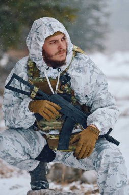 Beyaz kar kamuflajı ve taktik yelek giyen bir asker, soğuk hava operasyonları ve kış devriyesi sırasında etrafı incelerken elinde bir saldırı tüfeğiyle karlı bir ormanda diz çöker..